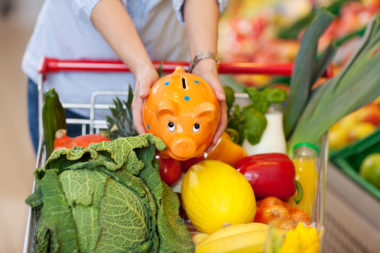 Woman holding an orange piggy bank with blue polka dots over her cart full of brightly colored produce.
