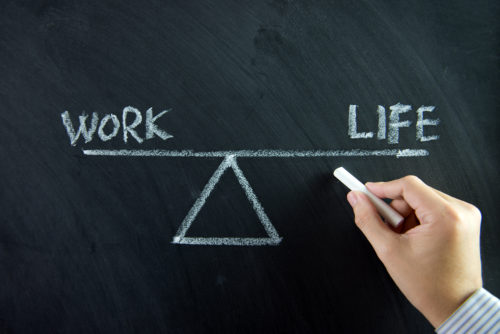 A hand drawing a scale on a chalkboard with the words "work" and "life" balanced equally on either end.