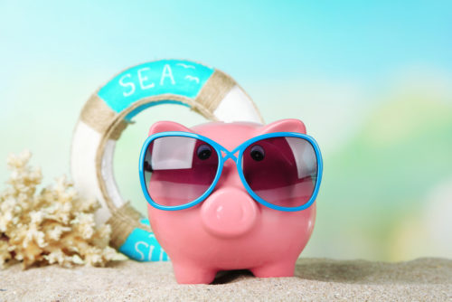 A piggy bank standing in the sand, wearing sunglasses with a life preserver behind it.