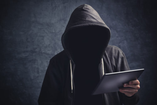 A cyber criminal with a hood hiding his face holds a tablet in his hand