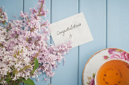 A simple note reading "Congratulations" in cursive lying on a table between flowers and a cup of tea