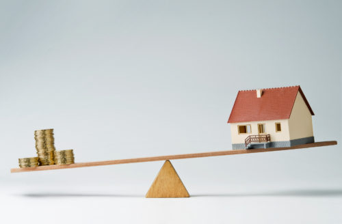 A model house and stacks of coins balancing against each other on a seesaw