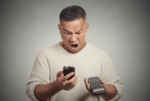 A man stares at his phone in open-mouthed shock while holding a calculator