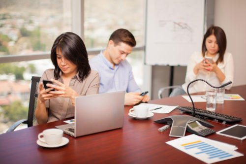 Three people at a conference table are distracted from work by their phones