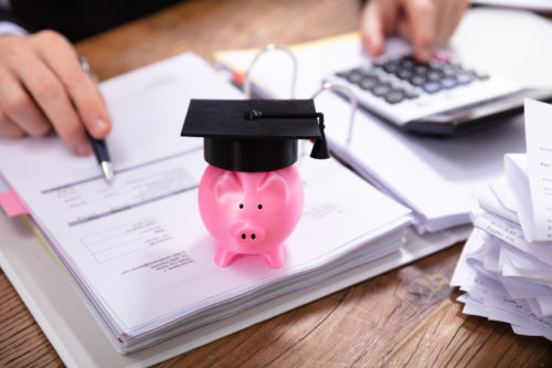 A pink piggy bank wearing a graduation mortar board, sitting atop a 3-ring binder, near a pile of receipts and a calculator
