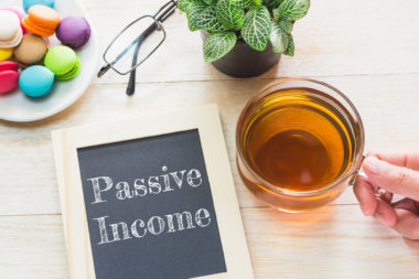A table has some cookies, a plant, eyeglasses, a cup of tea, and a chalkboard that reads "passive income" on top of it.