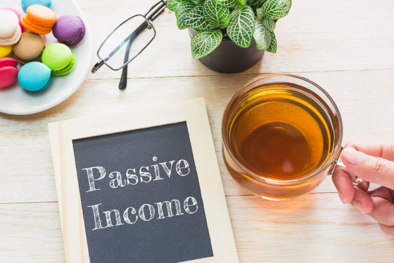 A table has some cookies, a plant, eyeglasses, a cup of tea, and a chalkboard that reads "passive income" on top of it.