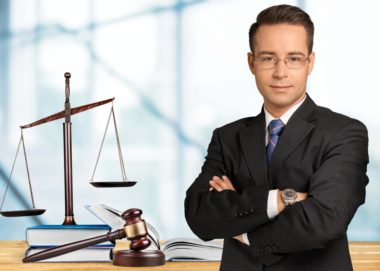 A lawyer stands crossed-armed next to a gavel and a balance scale.