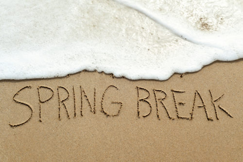 The words "Spring Break" are written in the sand on a beach with water washing up on it.