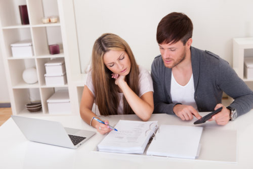 A couple sitting at a desk at their home, going over their personal finances via a computer and notebook.