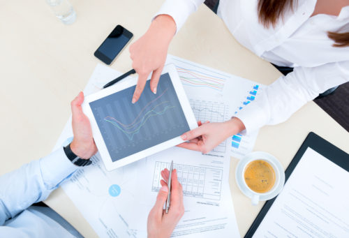 A financial analyst shows a man a tablet displaying a graph of an investment strategy with documents containing graphs of more strategies.