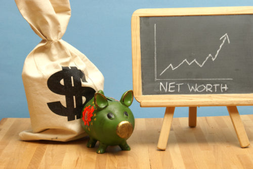 A piggy bank and a bag with a dollar sign on it sit next to a chalkboard showing a net worth graph with an arrow trending upward.