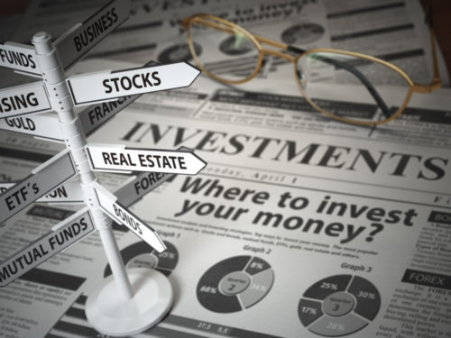 Eyeglasses sit on top of a newspaper that reads "Where to invest your money?" While a graphic of a direction sign has pointers on it with diverse investment options including "Business," "Real Estate," "Mutual Funds," "Bonds," and more.