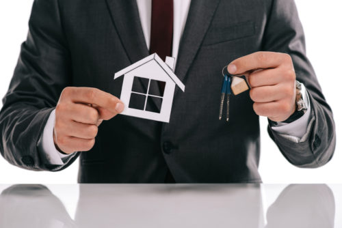 A mortgage broker holds a tiny model house in one hand and keys in the other.