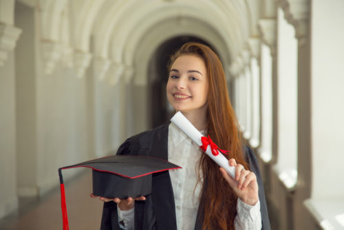 A young potential businesswoman wearing a gown holds her graduation cap in one hand and her business degree in the other.