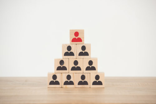 Blocks with black figures resembling people on them are put together in a pyramid fashion with the top figure being red, symbolizing the hierarchy of a pyramid scheme.