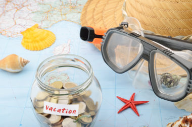 A pair of swimming goggles, a straw hat, and a jar marked "vacation" with change in it sits on top of a map.