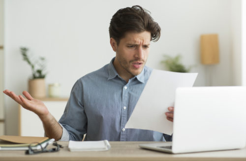A frustrated millennial looking over a financial statement at his desk where a computer sits.