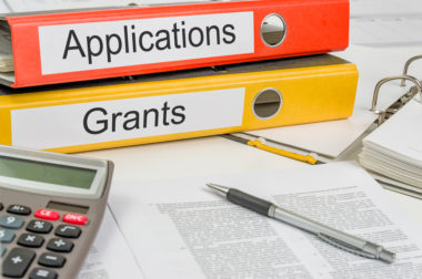 Documents, a calculator, a pen, and notebooks labeled "applications" and "grants" sit on top of a table.