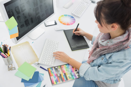 An artist sits at a desk, drawing on a pad connected to a computer with other art supplies scattered around.