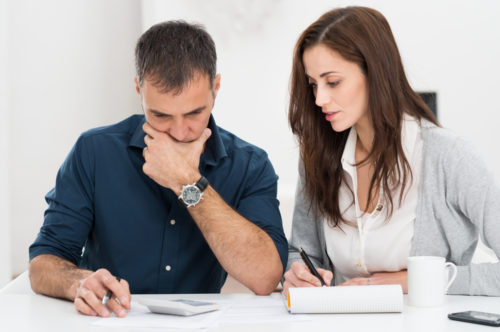 A couple sitting down at a table, calculating their budget while one writes in a notepad and the other types on a calculator.