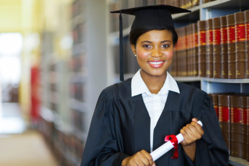 A woman in a cap and gown, holding her law degree next to some legal books.