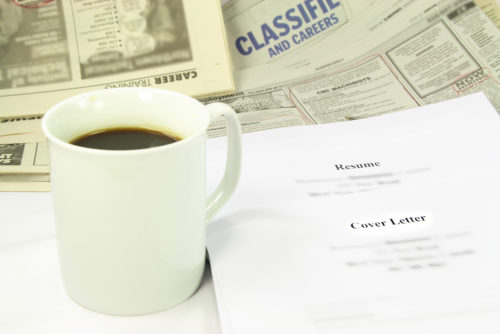A cup of coffee sits on a table along with the classified section of a newspaper, a resume, and a cover letter.