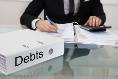 A financial advisor writes in a notebook while typing on a calculator with another notebook in front of him labeled "debts."