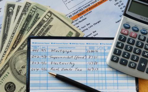 A checkbook sits along some cash and a calculator balanced with the words "mortgage," "supermarket (food)," electricity," and "real estate tax."