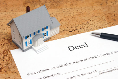A tiny model house and a pen sit on top of a document labeled "deed."
