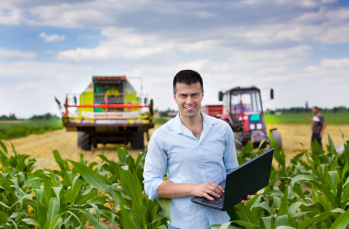 An agricultural engineer stands in a field with a laptop with farming equipment in the background.