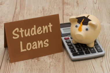 A piggy bank with a graduation cap sits on top of a calculator while next to a sign that reads "student loans."