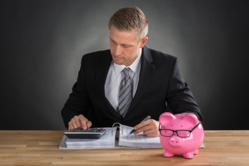 A real estate agent calculated 401(k) figures on a desk with a piggy bank in front of him to see if his client can buy a house.