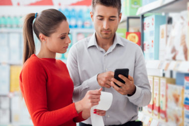 A couple checking a receipt at the supermarket while looking at their budget on a smartphone.
