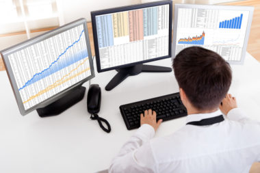 A man buying and trading stocks at his desk with three computer screens.