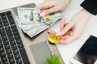 A woman sitting next to her laptop on a desk, holding cash in one hand and a bitcoin in the other.