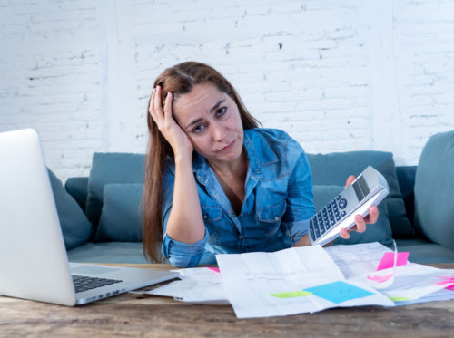 A frustrated woman holding a calculator with a mountain of debt papers and a laptop on her table.