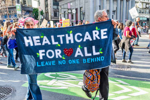 Participants of an event march down the street holding a banner that reads "Healthcare for all - leave no one behind."