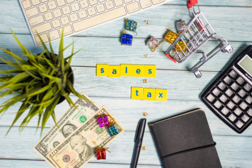 Tiles that spell out the words "sales tax" are surrounded by a notebook and pen, calculator, cash, a plant, a computer keyboard, and a tiny model shopping cart.
