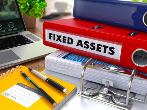 A table is cluttered with a laptop, pens, and notebooks — one of them labeled "fixed assets."