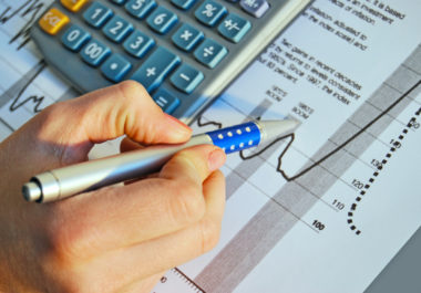 A hand holding a pen points to investment documents on a desk next to a calculator.