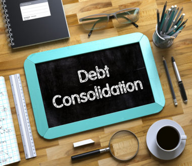 A chalkboard that reads "debt consolidation" lays on a table among a magnifying glass, a notebook, eyeglasses, and pens.