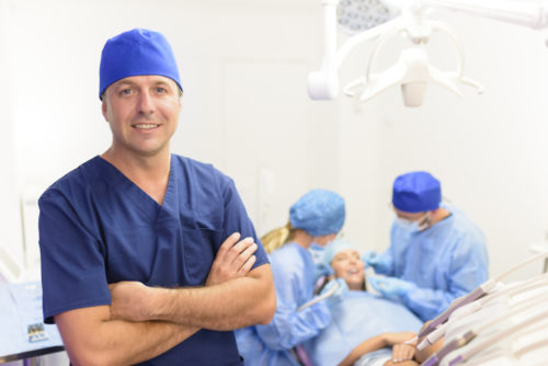 A maxillofacial surgeon smiling at the camera while his colleagues operate in the background.