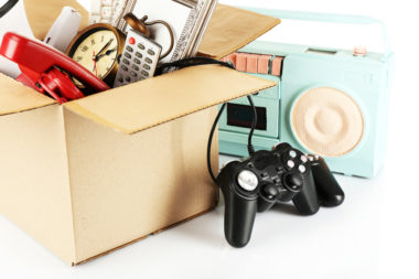 A box is filled with miscellaneous things that are ready to be sold, including a gaming controller, a clock, a radio, and more.