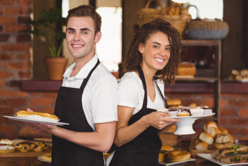 Two servers standing back to back, smiling while holding plates of food in a restaurant.