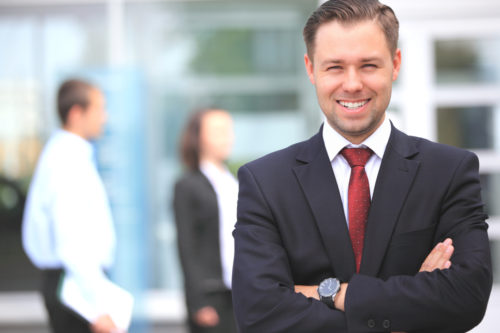 A banker stands, smiling at the camera, while his colleagues walk by in the background.