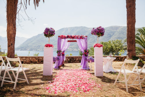 A pink wedding arch with flowers overlooks the sea and some mountains as part of a destination wedding.
