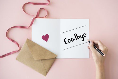 A woman writes "goodbye" in a card while an envelope and ribbon sits next to in on a desk.
