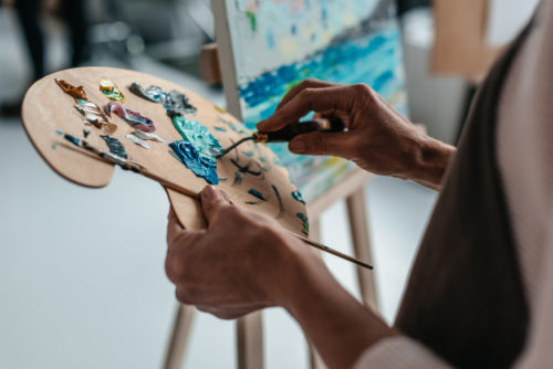 An artist holding a palette and paintbrush next to a canvas.