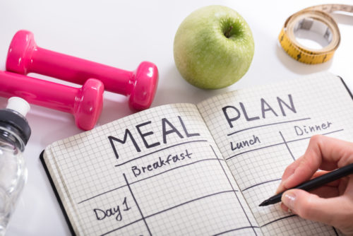 A person writing in a notebook on a table with the words "meal plan" written at the top, with weights a fruits.